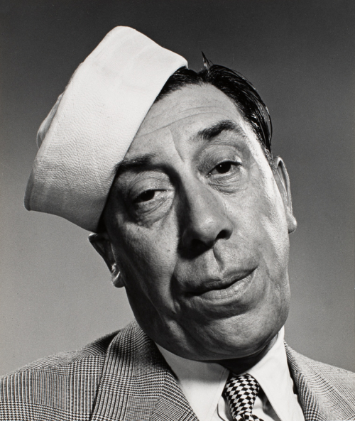 Head-shot of man in plaid jacket and checked-tie. His head is tilted to the left and he wears a sailor's cap   