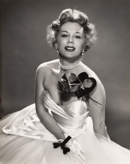 Woman in strapless white dress leaning forward, towards viewer. Wearing satin gloves, black bracelet; a large corsage