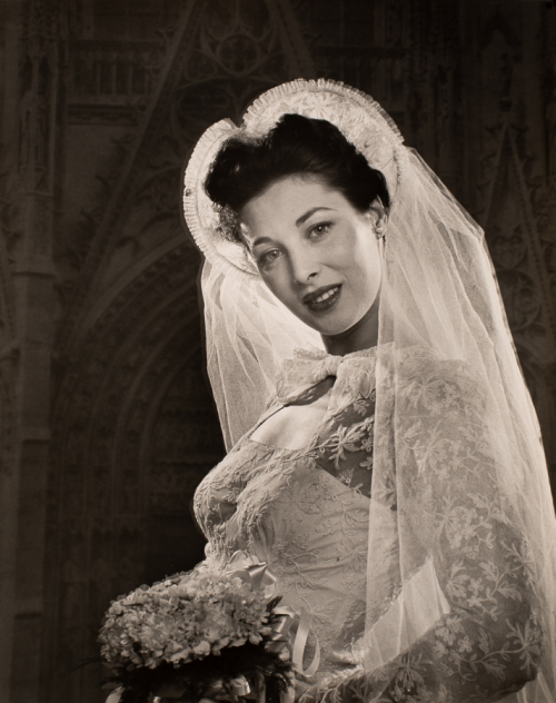 Woman wearing wedding gown and long veil, facing left side of image with head turned towards the viewer and tilted