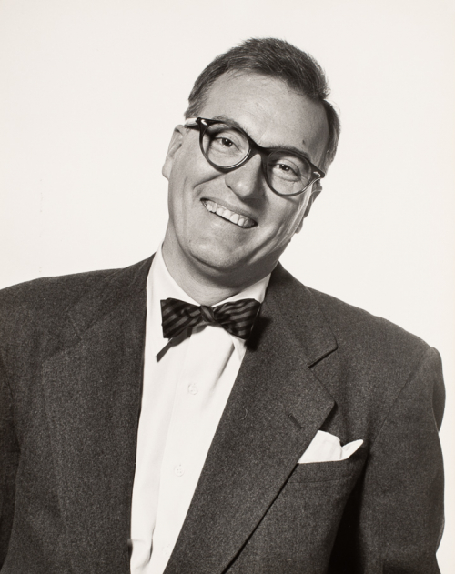 Man facing viewer, head tilted towards the right side of image and smiling, wearing dark suit, bowtie, and glasses