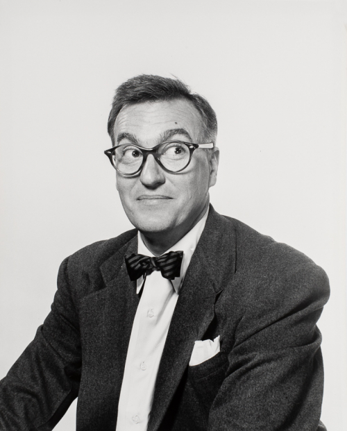 Man in a suit and bowtie and glasses shown from waist up, body tiled to left head facing viewer glancing up-right