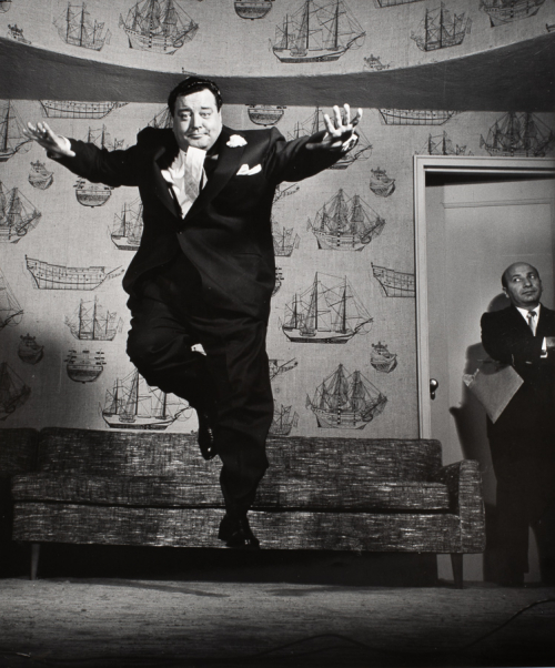 Man in mid-jump in what looks like a living room scene, to right of image man at slightly opened door arms crossed