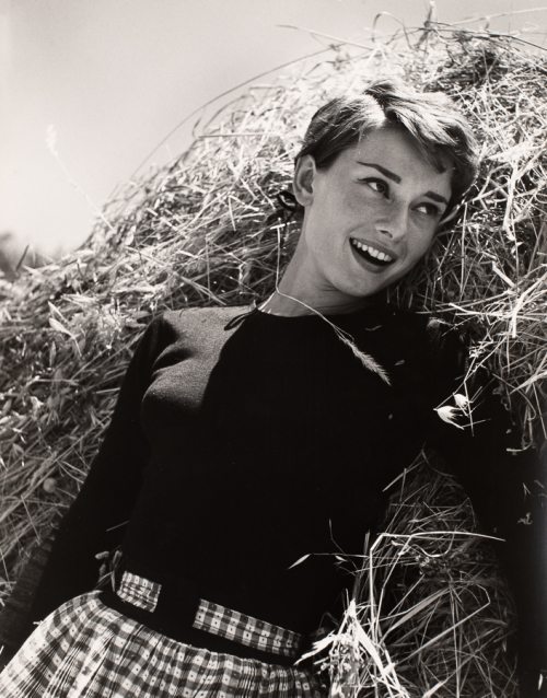 Short dark haired woman from hips up wearing black top and checked bottoms leaning on hay on right of image