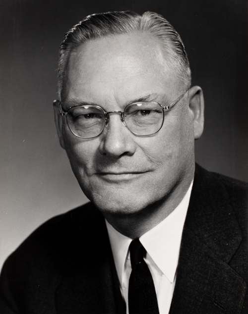 Bust-shot of man wearing wire-rimmed glasses and dark suit and tie, turned slightly towards the left