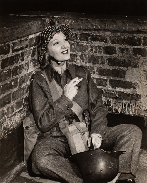 Woman seated smoking a cigarette near brick wall dressed in military uniform with canteen around her neck and helmet at her feet