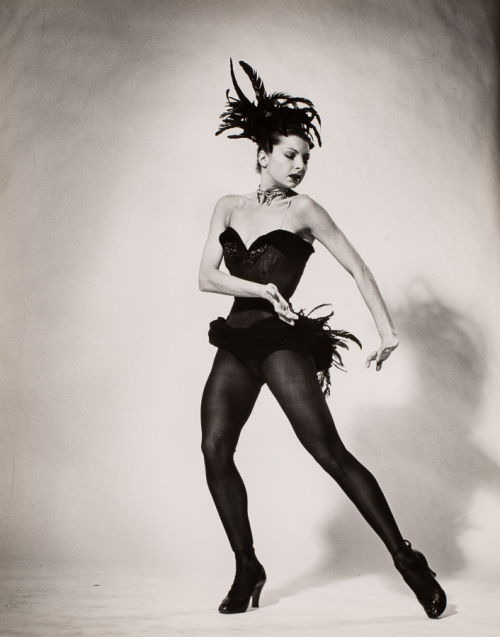 Woman dressed in black leotard, nylons, and feathered bustle and hat. Body facing forward left arm, leg, head are turned right
