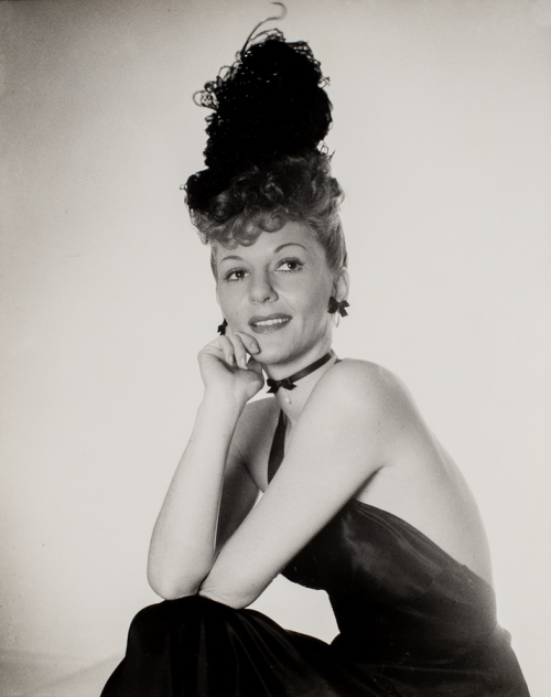 Woman in black dress seated with elbows resting on knees and chin resting on right hand. Hair up in a dark feathered piece