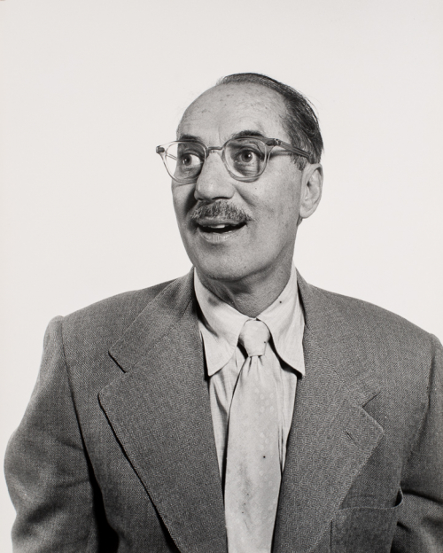 Bust-shot of man wearing tweed jacket, linen shirt and tie, and glasses facing viewer but with head to left