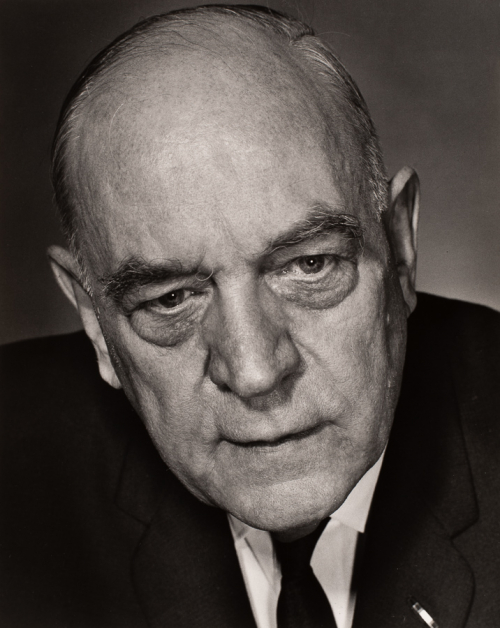 Head-shot of older man facing viewer with head tilted towards left side of image. He is wearing a dark suit and tie