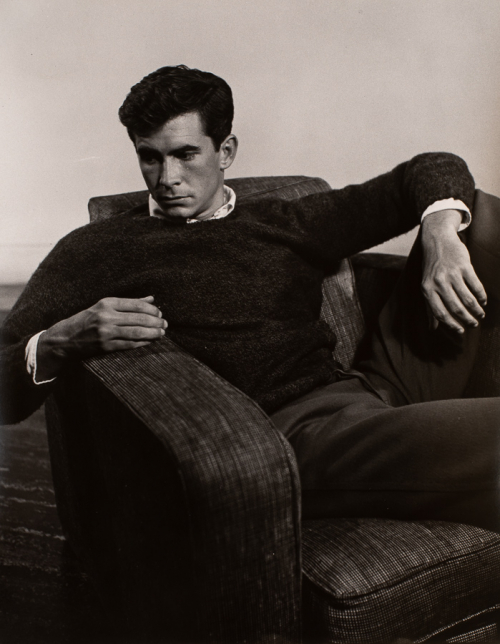 Man sitting in dark upholstered chair. Wearing dark trousers and a sweater. Left leg is over arm rest, left arm is on left leg 