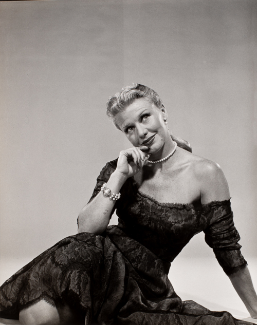 Woman seated on floor, facing viewer and leaning on her left hip and arm and towards right side of image. Wearing a dark dress