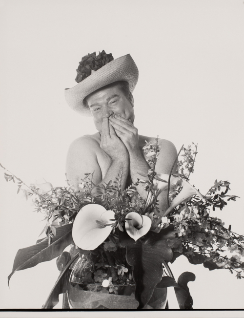 Man behind large vase of flowers in straw hat with flowers on it appears to be shirtless with hands covering mouth bashfully