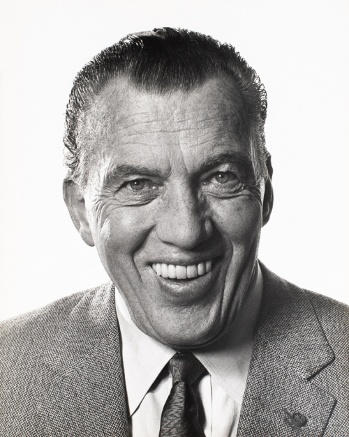 Head-shot of man in medium-grey jacket and tie with head turned slightly towards right side of image and smiling.