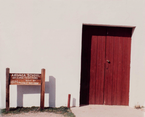  exterior of a white painted building. Prominent to the right is a red wooden door, and to the left is a wooden sign with white 