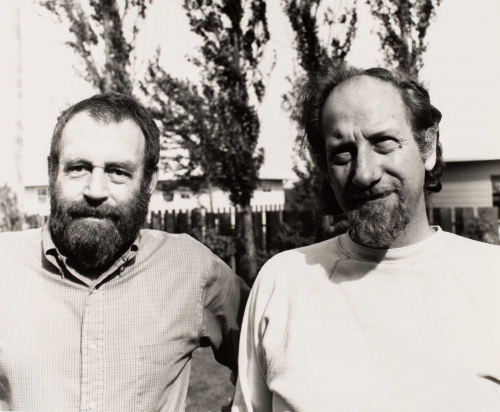 A portrait of two artists, Ralph Haskell (left) and David Delafield (right), standing outdoors with fencing, trees, and houses 