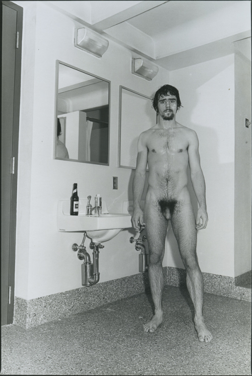 Black and white face-frontal full-length photograph of a naked man in a public restroom