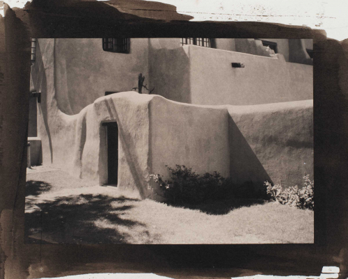 View of adobe house.  Door at oblique angle on left.  Bushes in center of image extending to right side.  Shadow of tree in LL