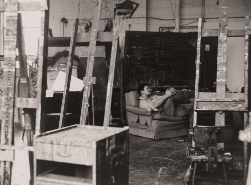 black and white image blackboard in background; man lounging on a couch; easels all around