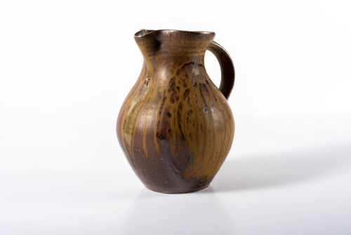 A side view of a small gourd shaped pitcher glazed in brown with caramel-colored drips.