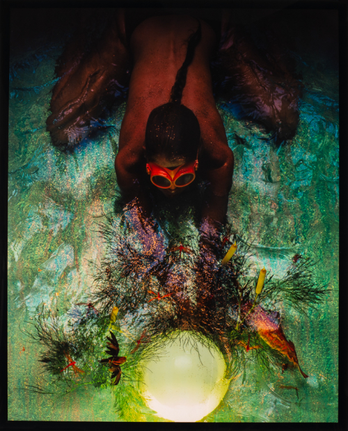 overhead view of a figure with braided hair, wearing red goggles crouching in shallow water reaching for glowing sphere