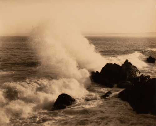 Water is in the lower 3/4 of the image; distant land and sky are along the upper edge; waves crash against rock
