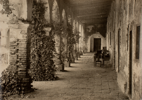 Crumbling wall with doorways and windows along right side receding into picture plane to the left.  Stone walkway with chair 