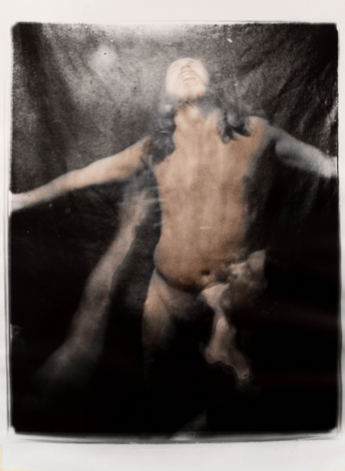 Nude long-haired man with arms outstretched looking up a light or water coming down from top, blurry person and arm