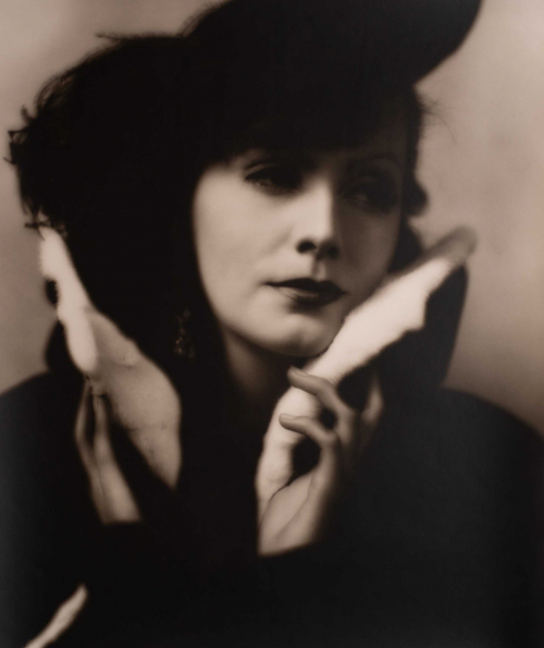 soft focus image of woman looking to right, wearing dark-plumed hat. Her hands turning up the light fur-trimmed coat collar