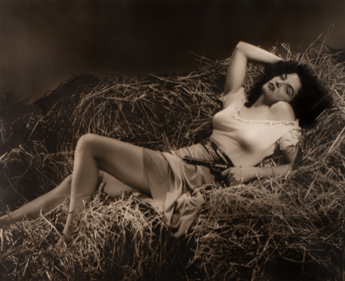 Woman lying on pile of straw, her right arm is above her head and  holding a revolver in the other hand at her hip
