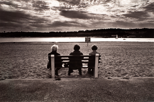 Rear-view of 3 women on bench below center of image. Beach in front of them, with water and boats beyond beach. 