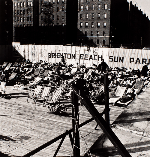 Fully-clothed people lying on lounges facing LL. 'Brighton Beach Sun Park' painted on fence behind 