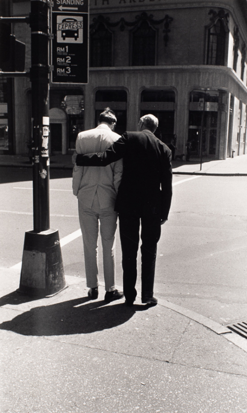 Rear view of two men standing on street corner. Man on left is wearing light suit and right is wearing dark suit.