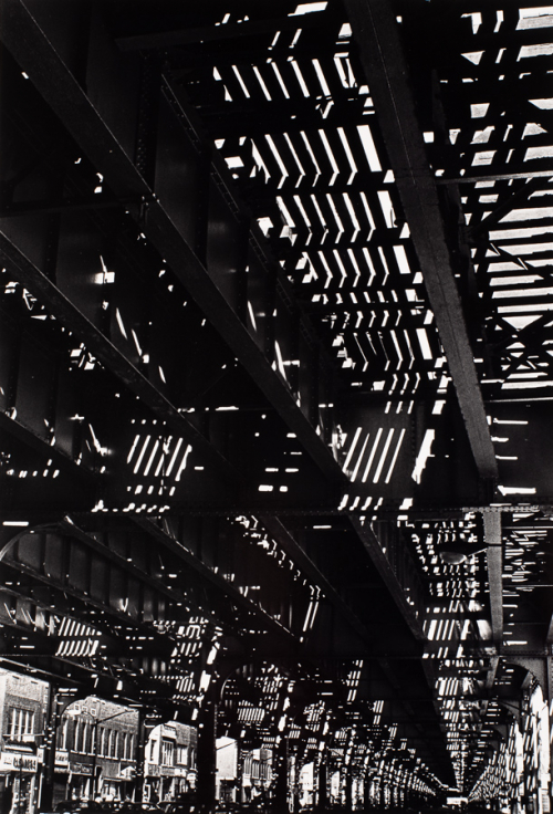 View under structure of elevated train. Sunlight filtering through tracks. Store fronts and parked cars visible along lower edge
