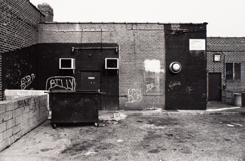 View from behind a brick building; concrete blocks and garbage dumpster in LL quadrant,  graffiti painted on walls behind