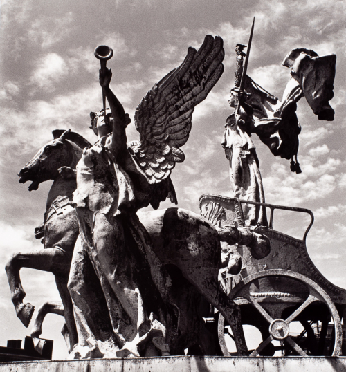 Sculpture of winged trumpet player facing viewer.  Horse and chariot with Female figure with sword riding. Cloud filled sky.