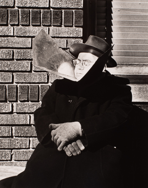 Man dressed in dark coat and hat seated on sidewalk leaning against brick wall. Has reflective sunning device under his chin 