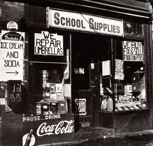 View of storefront from oblique angle. In window large sign reads "School Supplies" with several other signs and objects.