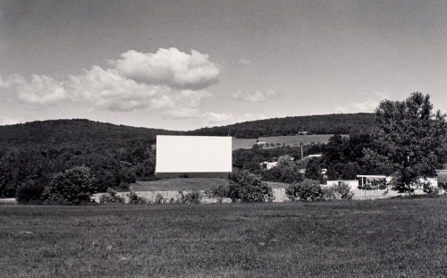 Grass in lower third, rectangular movie screen left of center in middle zone; wooded hills and partly cloudy sky in upper third