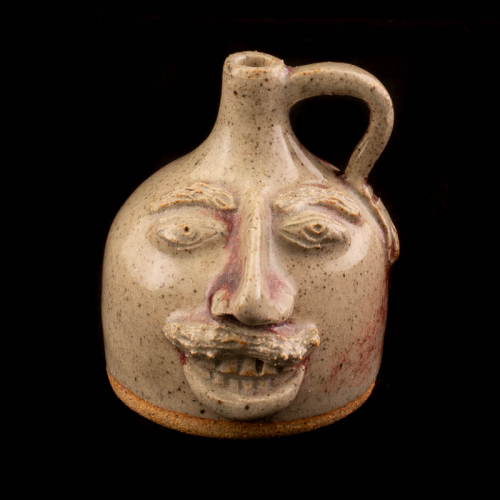  tiny face mug with a shiny biege glaze, a right side handle, a prominent nose and mustache, and a smile showing upper and lower