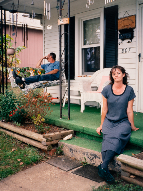 A porch of a white house with black shutters. A woman sitting on green carpeted porch steps, man lounging in back