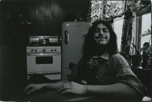Portrait of a seated man with long hair, grinning with a cigarette in his mouth; in a kitchen