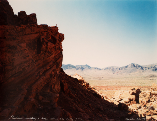 Rocky landscape with high, shadowed ledge in left half and a person seen on top. Clear, blue sky in upper half of image 