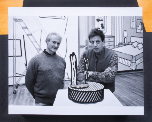  black and white photographic image of two males flanking a small sculpture on a pedestal top within an art gallery setting