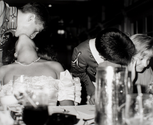Two Couples at table-one male in uniform at left bending over and kissing seated female whose head is thrown back. 