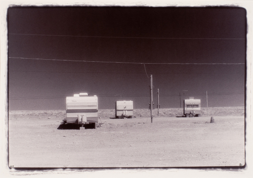 Sand or gravel-covered landscape in lower, dark sky and wires above. Three campers one in front left two further back