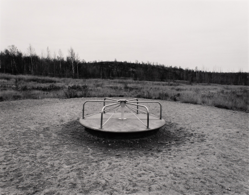 Empty merry-go-round in center of image surrounded by sand /gravel.  Tall grass, trees, and light sky in background