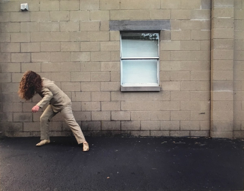 A figure wearing beige poses in front of a beige cinder block wall with a window