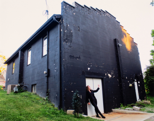 A female figure dressed in black poses in front of a white door at the back of a black-painted building