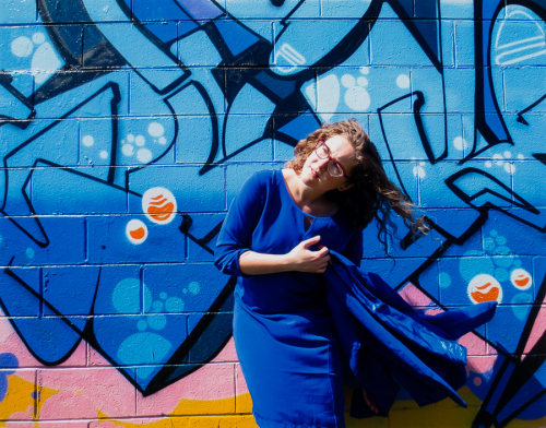 An image of a female dressed in blue standing in front of a wall of graffiti, which is primarily blue