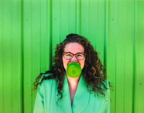The torso and face of a female who wears a green jacket, bites a green apple, and is in front of a green wall
